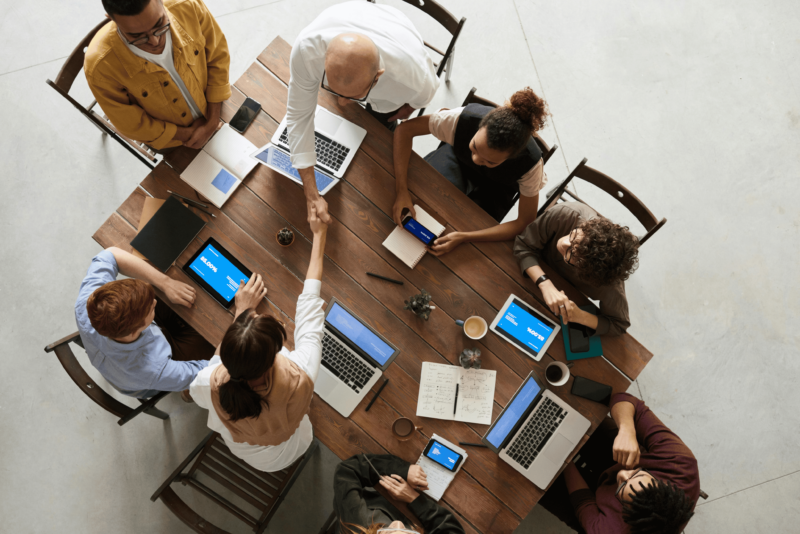 A group of people sitting around a table with computers and coffee. Two are shaking hands.